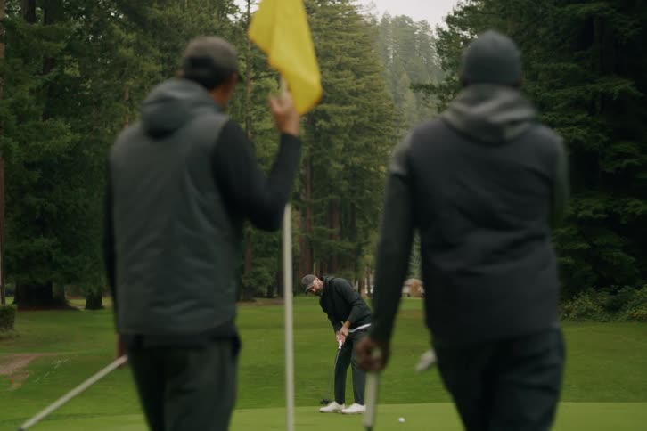 A man sinks a putt on the green as his friends watch on a beautiful, tree-lined golf course