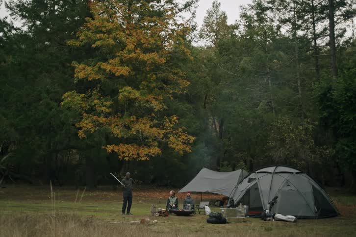 A group of three male friends relax around a campfire at their rustic campsite in a forest clearing during autumn