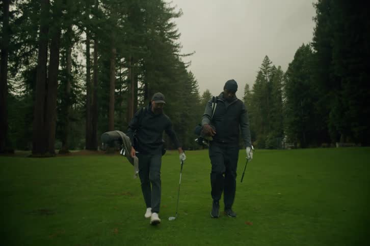 Two male golfers walk in slow motion across a lush green fairway surrounded by tall evergreen trees