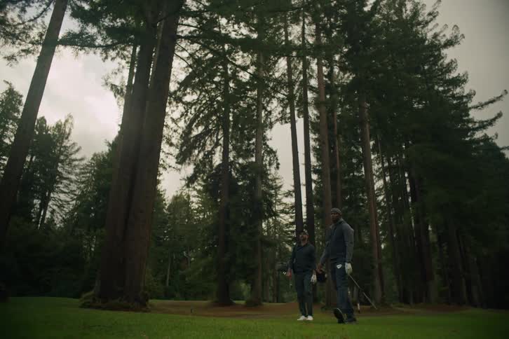 A golfer takes a powerful swing in slow motion on a beautiful course surrounded by towering redwood trees while his friend watches