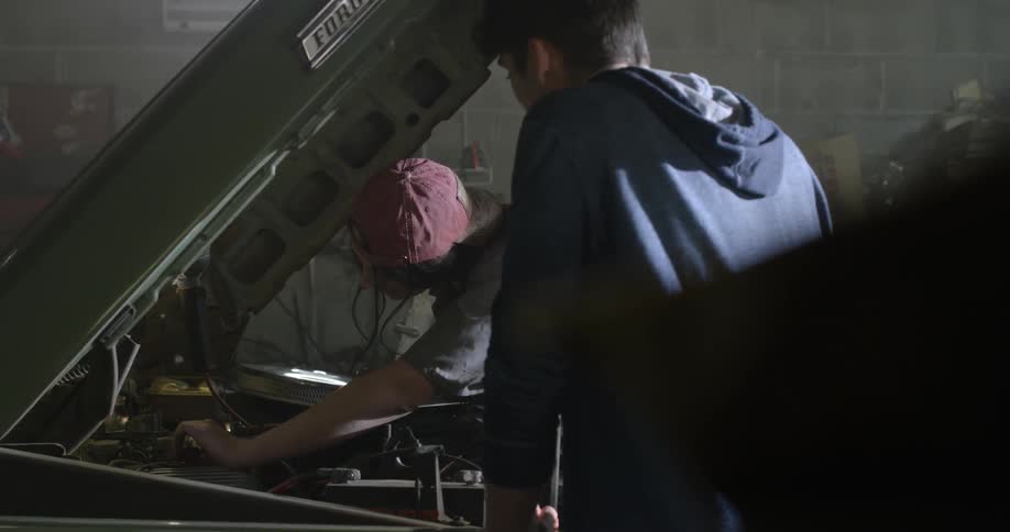 A woman is working on a car engine as a man talks to her