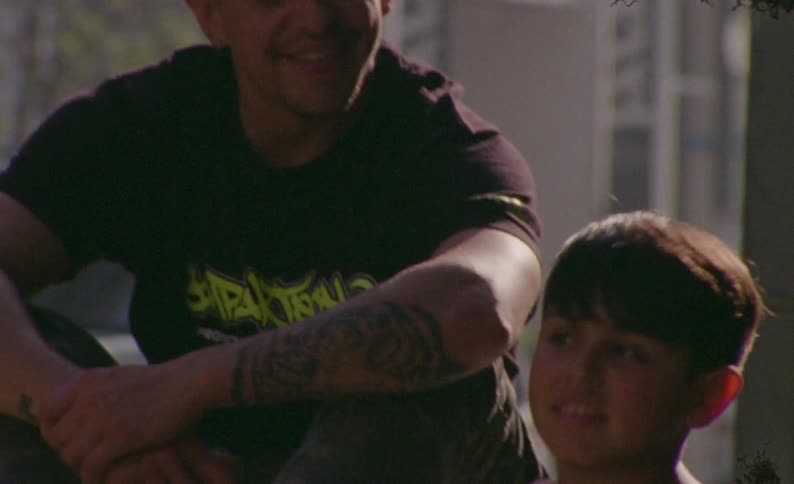 A tattooed father wearing a beanie smiles and talks with his young son outdoors