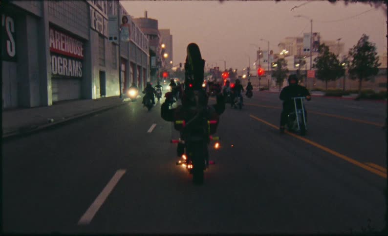 A motorcyclist performs a wheelie while riding with a large group of bikers down a city street at dusk in slow motion