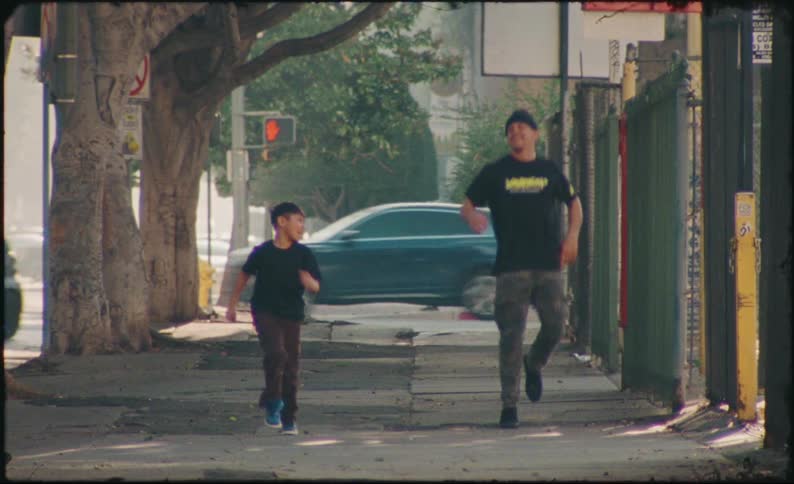 A happy father and son joyfully run, jump, and skip down a city sidewalk together in this vintage-style slow motion clip