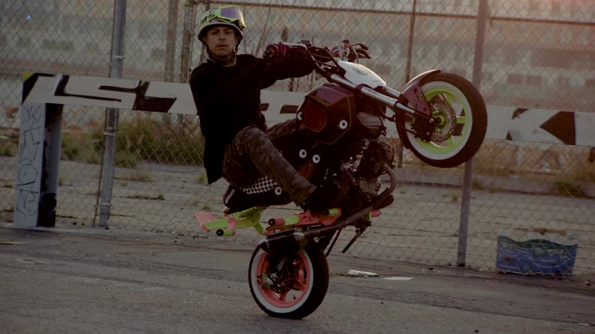 A talented stunt rider skillfully balances a colorful custom motorcycle on its back wheel in an urban industrial lot at sunset
