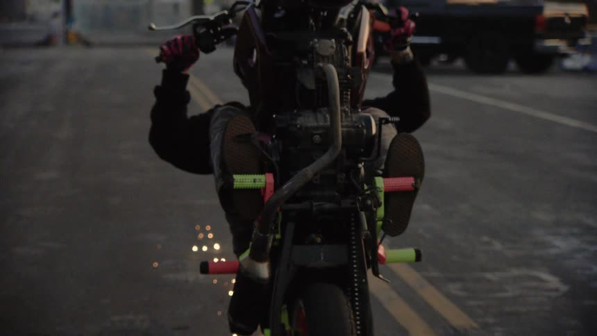 A stunt rider performs a wheelie on a motorcycle down a street, creating sparks in a dramatic slow-motion shot from behind