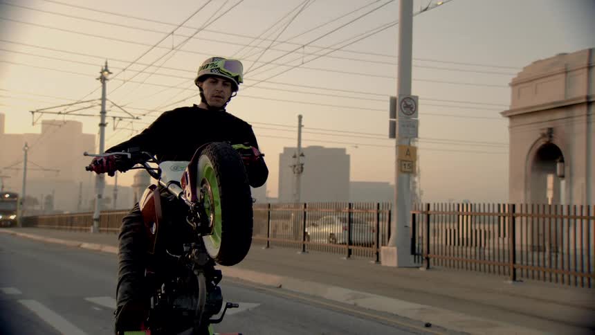 A motorcyclist performs a wheelie stunt in slow motion on a city bridge during a hazy sunset