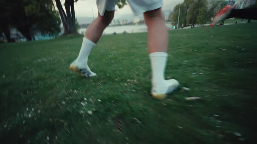 A dynamic, low-angle tracking shot follows a soccer player's feet in slow motion as they run and dribble the ball across a grassy park