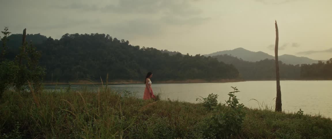 A solitary woman stands peacefully on a grassy bank, looking out over a serene mountain lake at golden hour
