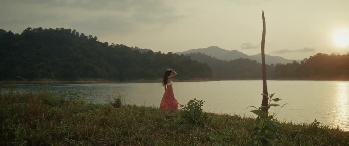 A graceful woman in a flowing skirt walks along a serene lakeside, pausing to enjoy the beautiful sunset over the distant mountains