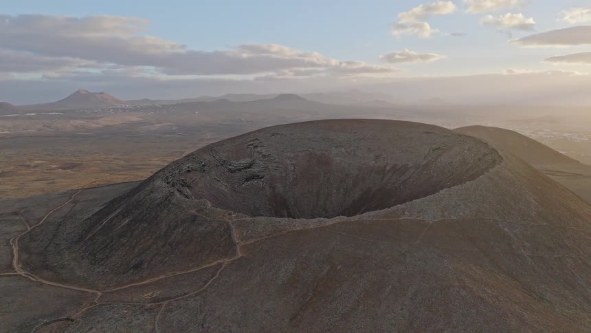 A slow-moving aerial shot captures the immense scale of the Calderón Hondo volcano crater during a hazy golden hour