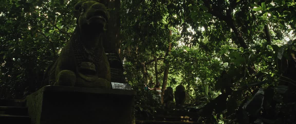 Travelers walk up ancient mossy stone steps past a weathered statue in a lush green jungle