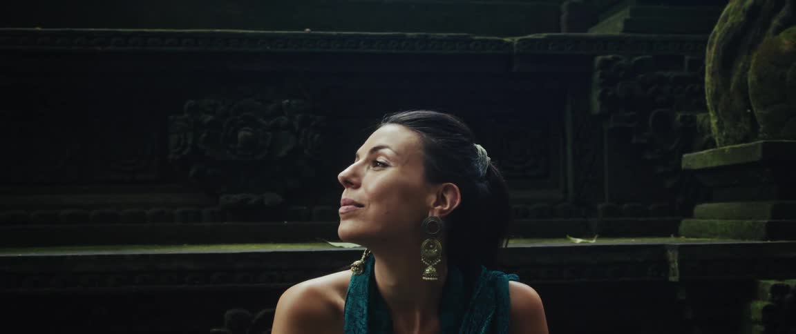 A serene woman gazes up in awe while sitting amidst the ancient, moss-covered ruins of a stone temple