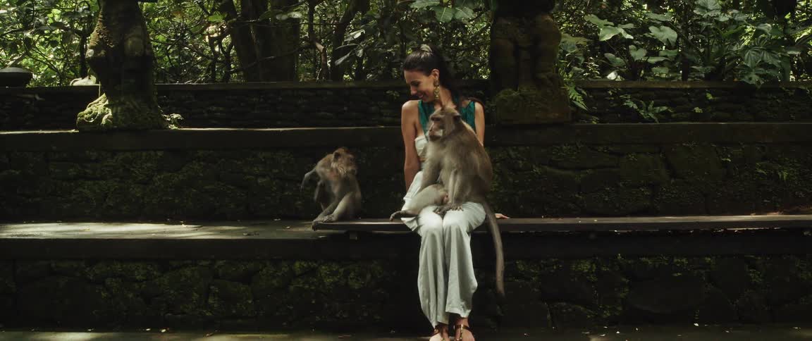 A smiling woman sits peacefully on a mossy stone bench in a lush forest sanctuary with two monkeys, one resting comfortably on her lap