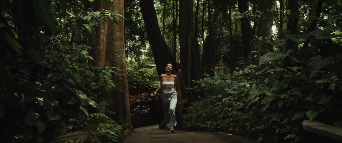 A woman smiles peacefully as she walks in slow motion along a stone path through a lush and dense tropical jungle