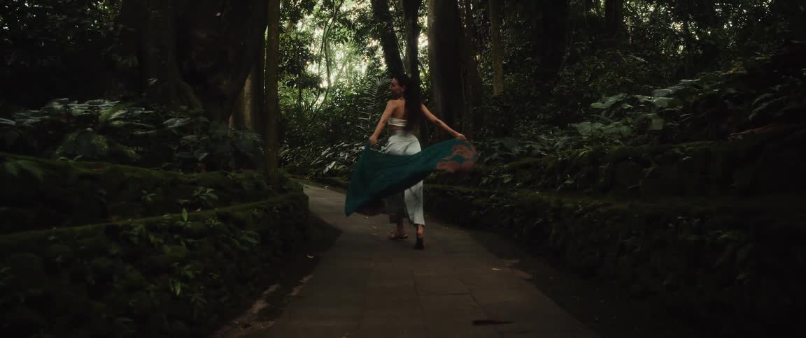 In slow motion, a carefree woman with a flowing teal scarf walks and smiles along a stone path through a lush, mystical green forest