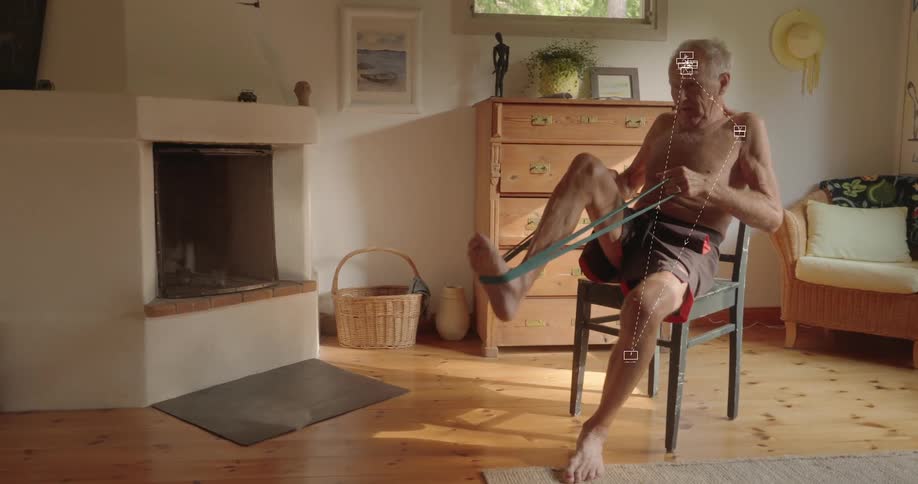 An elderly shirtless man sits on a chair in a sunlit room, performing leg exercises with a resistance band