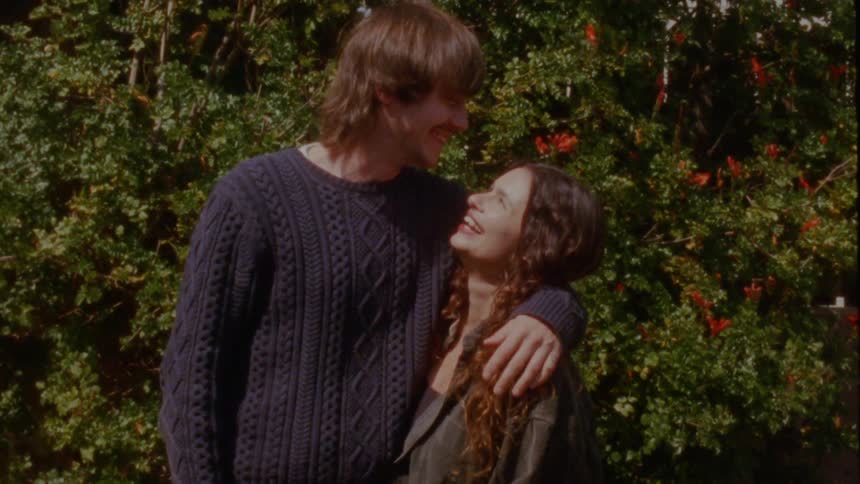 A happy couple laughs and embraces while standing in front of a flowering bush on a sunny day