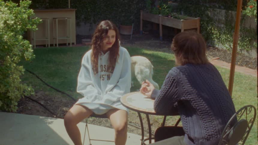 A young couple sits at a small table on a sunny backyard patio, engaged in a quiet conversation in this vintage-style footage