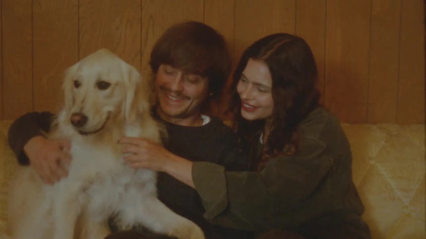 A happy young couple with a retro aesthetic lovingly pets their golden retriever while sitting on a couch