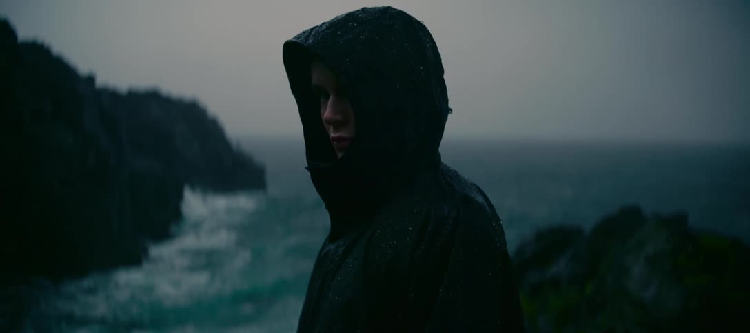 A lonely figure in a dark hooded raincoat contemplates the stormy sea from a cliff on a rainy, overcast day