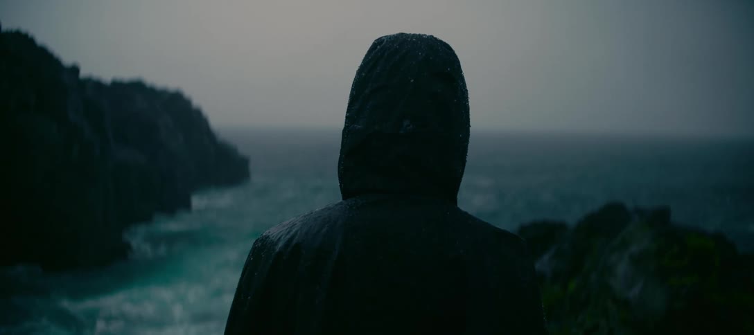 A solitary figure in a dark rain hood looks out over the choppy, dark teal ocean during a rainstorm
