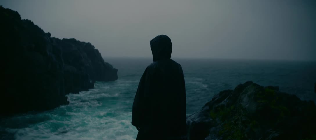 A solitary figure in a dark rain cloak contemplates the stormy sea from a rugged cliff on an overcast day