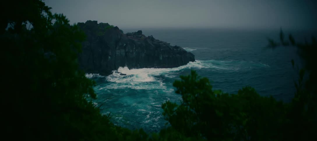 Viewed through dark foliage, powerful ocean waves crash against a dramatic rocky cliff on an overcast day