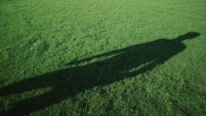 An elongated shadow of a person is cast across a vibrant, sunlit green lawn