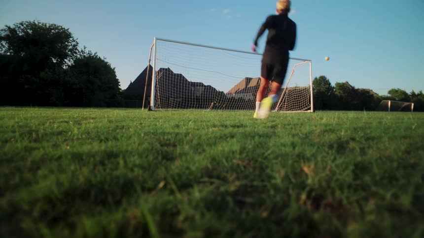 A soccer player scores a goal with a powerful kick in a dramatic slow-motion, low-angle shot on a grassy field