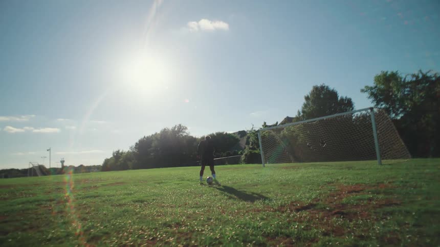 Slow motion footage from behind a soccer player as he juggles and dribbles the ball towards the goal on a sunny field