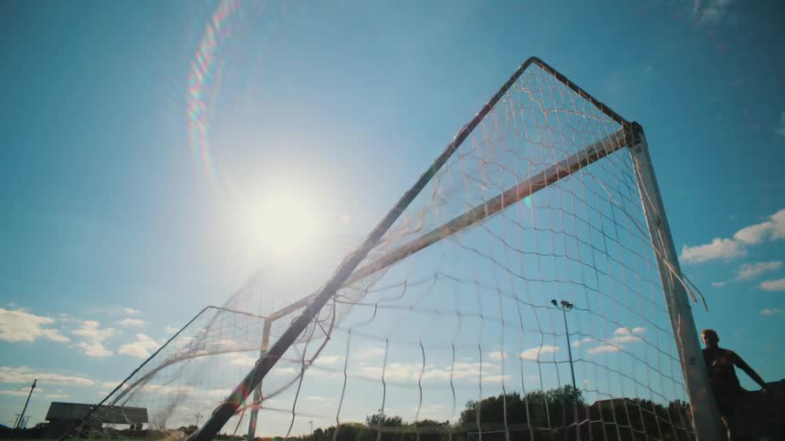 A soccer ball flies into the goal net in slow motion against the bright sun and blue sky