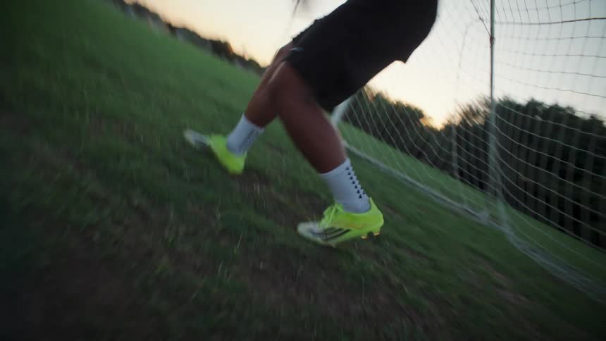 A dynamic, low-angle shot shows a soccer player kicking a ball into the goal on a grassy field at sunset