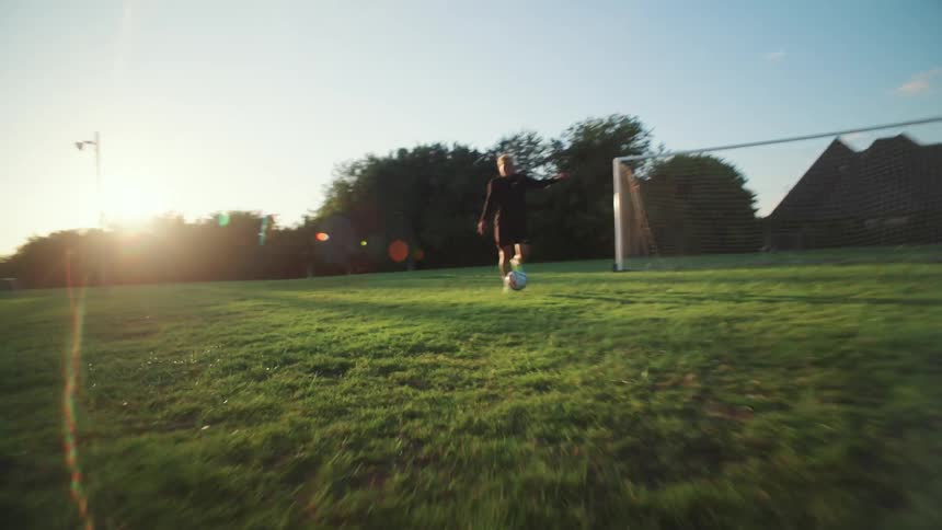 A male soccer player dribbles the ball down a grassy field at sunset in a dynamic low-angle shot