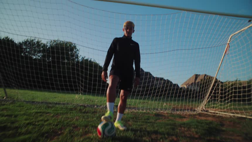 A low-angle shot reveals a confident young male soccer player with his foot on the ball in front of a goal