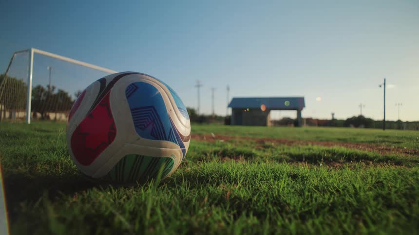 A vibrant soccer ball rests on a sun-drenched green field with the goal in the background