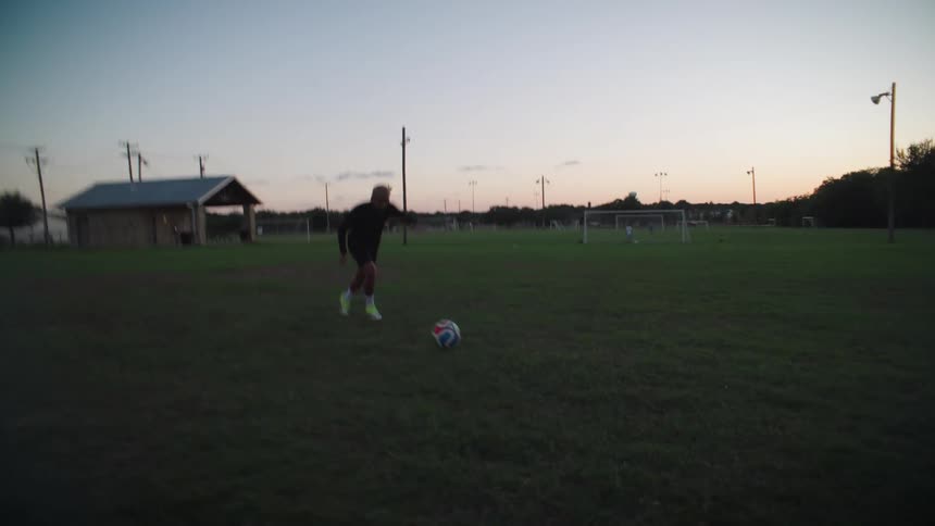 A male athlete skillfully controls a soccer ball before kicking it directly at the camera in dramatic slow motion on a grassy field at dusk