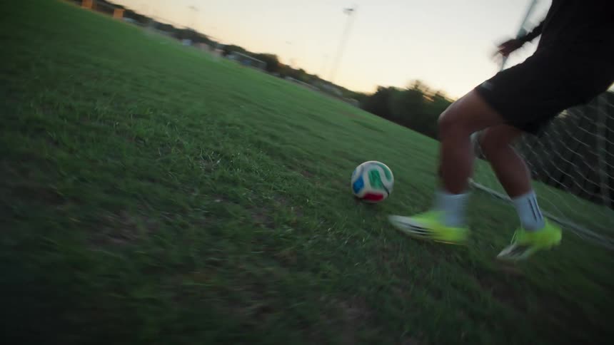 Dynamic low-angle slow-motion footage follows a soccer player kicking a ball on a grassy field at dusk