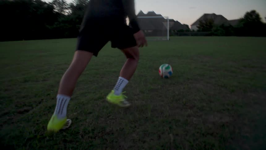 A soccer player skillfully controls a ball with his feet before taking a powerful shot toward the goal in a dramatic slow-motion sequence at dusk