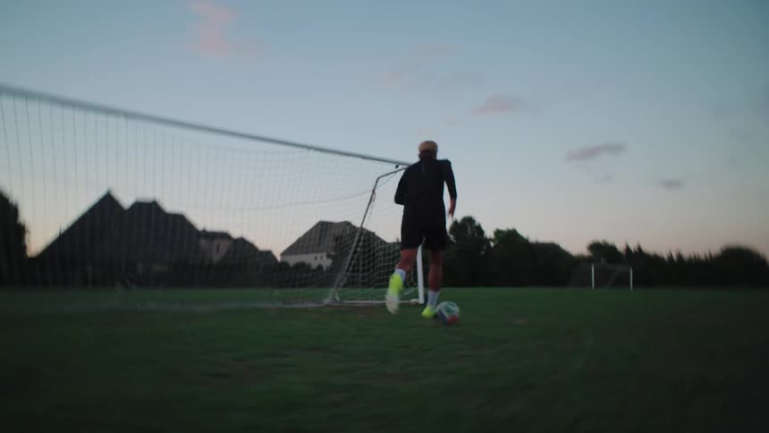 A young male soccer player practices his dribbling skills on a green field towards the goal during a beautiful dusk