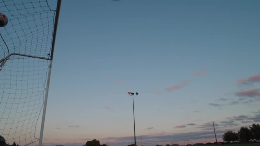 A soccer ball flies into the top corner of the goal in slow motion against a beautiful dusk sky