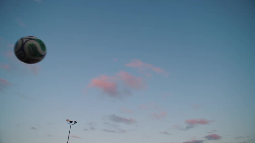 A soccer ball flies through the air in slow motion against a beautiful dusk sky with pink clouds