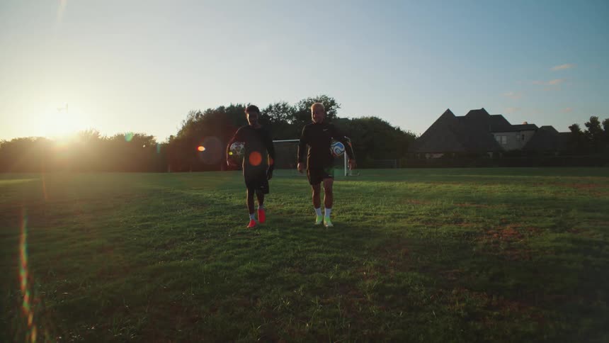 Two diverse young male soccer players walk towards the camera in slow motion across a field at sunrise