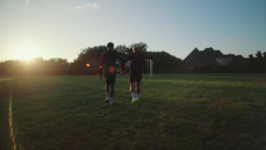 Two diverse male soccer players walk onto a green field with their soccer balls, ready to practice at sunset