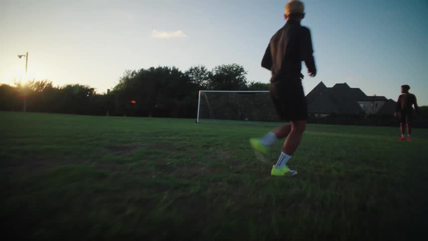 Dynamic point-of-view shot follows a soccer ball as one player passes to another who scores a goal on a field at sunset
