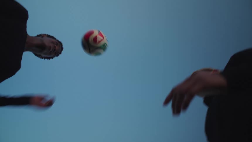 A worm's-eye view captures two diverse young men in slow motion as they skillfully head a soccer ball back and forth against the blue sky