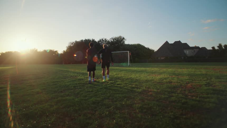 Two diverse male soccer players walk onto a green field towards the goal as the sun sets, ready for practice