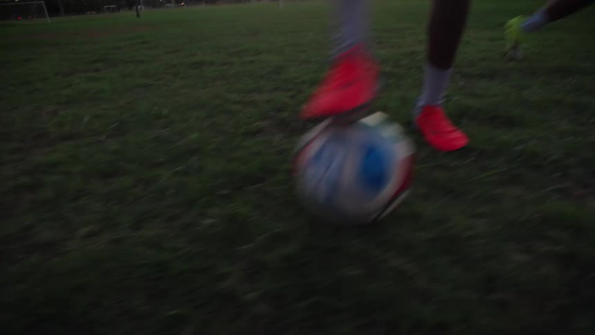 Dynamic slow motion footage tracks the feet of two soccer players as they compete one-on-one on a grassy field at sunset, ending with a goal