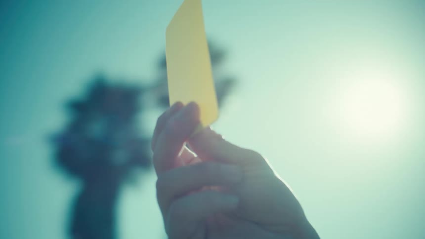 Slow motion close up of a hand holding up a yellow penalty card against a sunny sky with palm trees