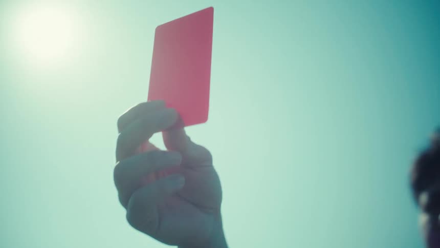 A referee holds up a red card against a sunny sky with palm trees in slow motion